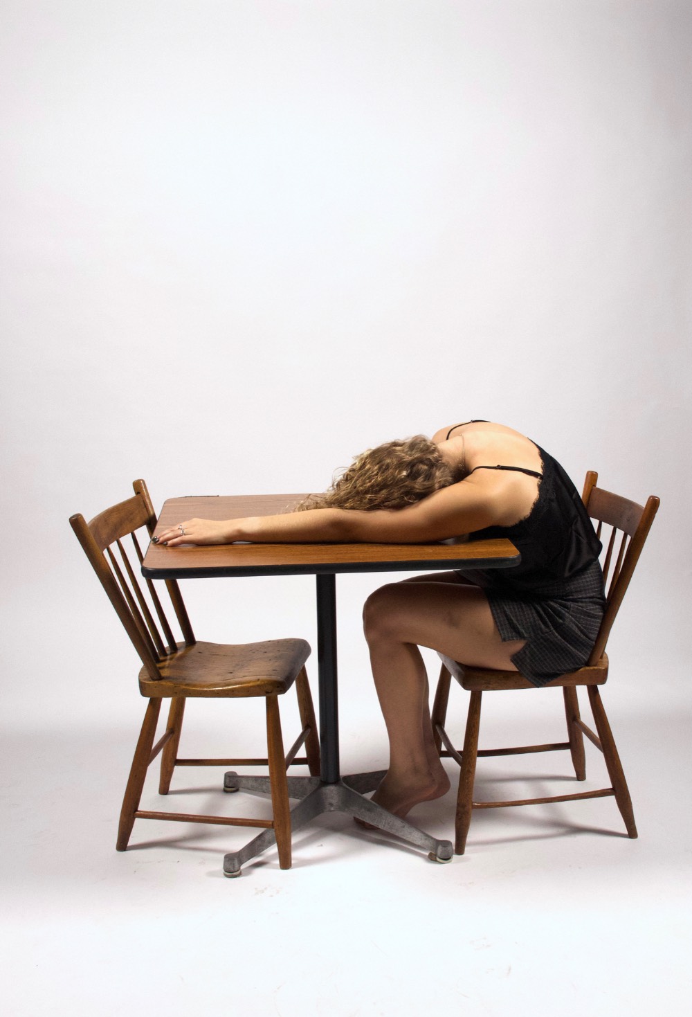 A side view of a woman in a black slip passed out on a table with two chairs.